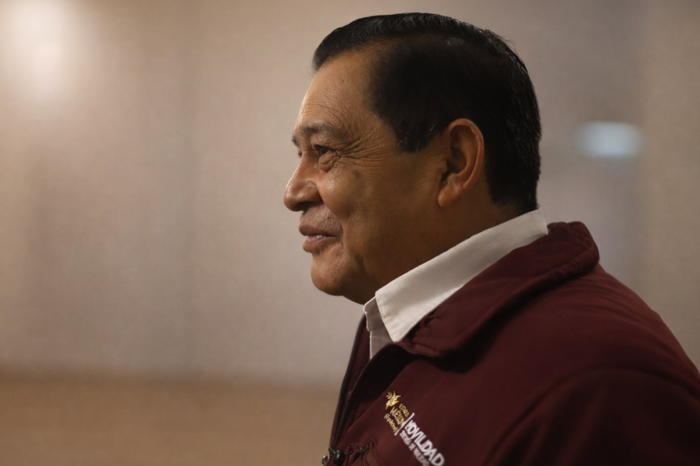Perfil de un hombre mayor sonriendo, vistiendo una chaqueta burdeos y una camisa blanca, en un entorno iluminado.
