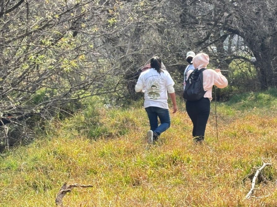 Tres personas caminando por un sendero natural, rodeadas de arbustos y hierba alta.