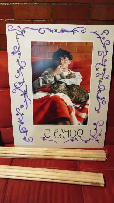 Cartel decorativo con una foto de un niño sentado en un sofá, acariciando a un gato. El nombre 'JESHUA' está escrito en la parte inferior, rodeado de un marco adornado con flores moradas.