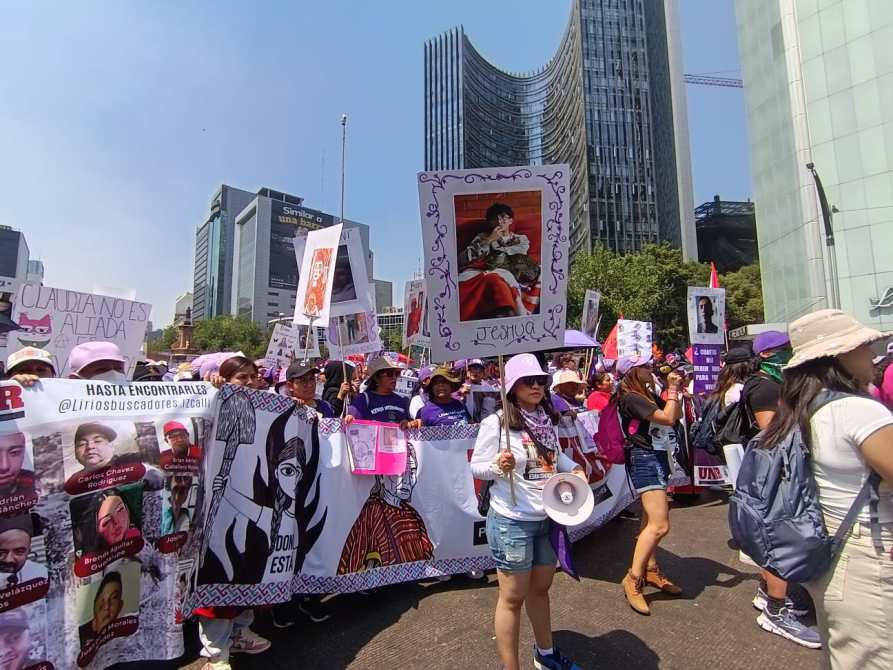 Manifestación con personas sosteniendo pancartas y carteles en un ambiente urbano, donde se observan sombreros de color púrpura y más de un diseño de carteles.
