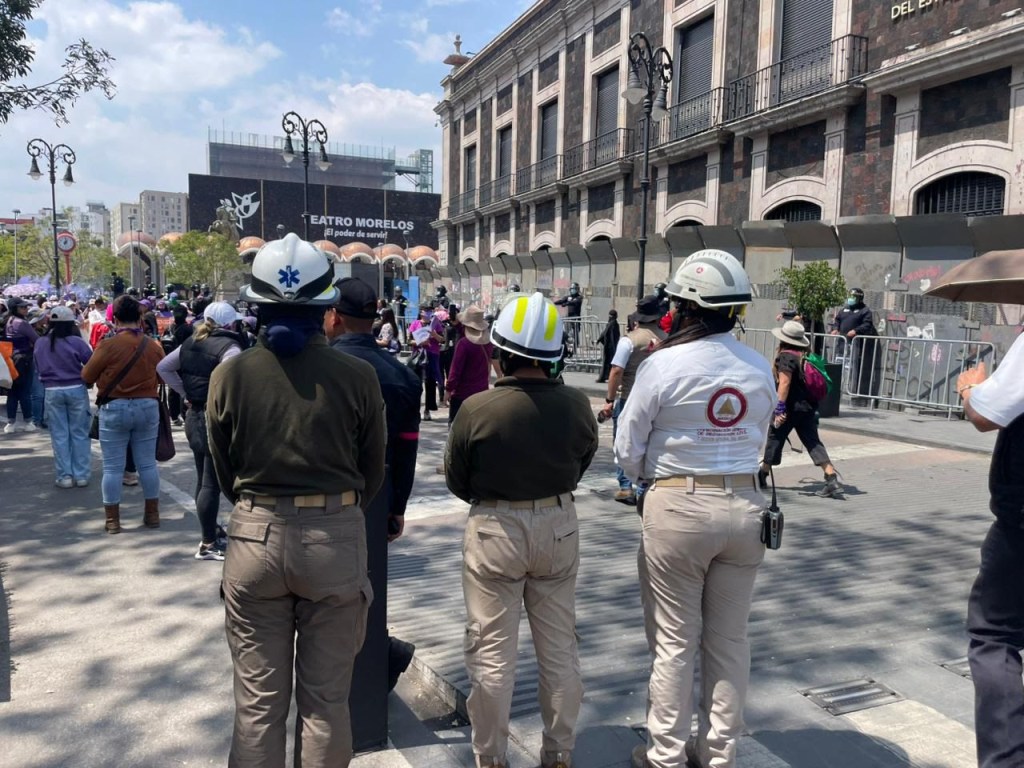 Tres personas de pie con cascos de seguridad observan a una multitud en una calle, con un edificio histórico al fondo.