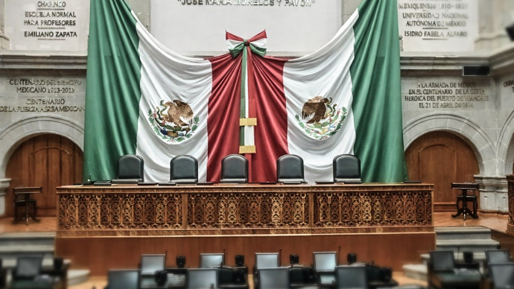 Escenario con banderas de México, en la sala del congreso. Tres sillas oscuras frente a un fondo decorado con el escudo nacional.