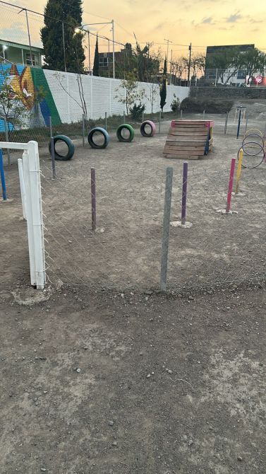 A playground area featuring rubber tires, colorful poles, and a wooden climbing structure, with a dirt surface and a fence.