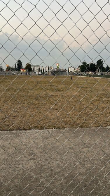 View of an empty, grassy field from behind a chain-link fence, with distant buildings and trees in the background.