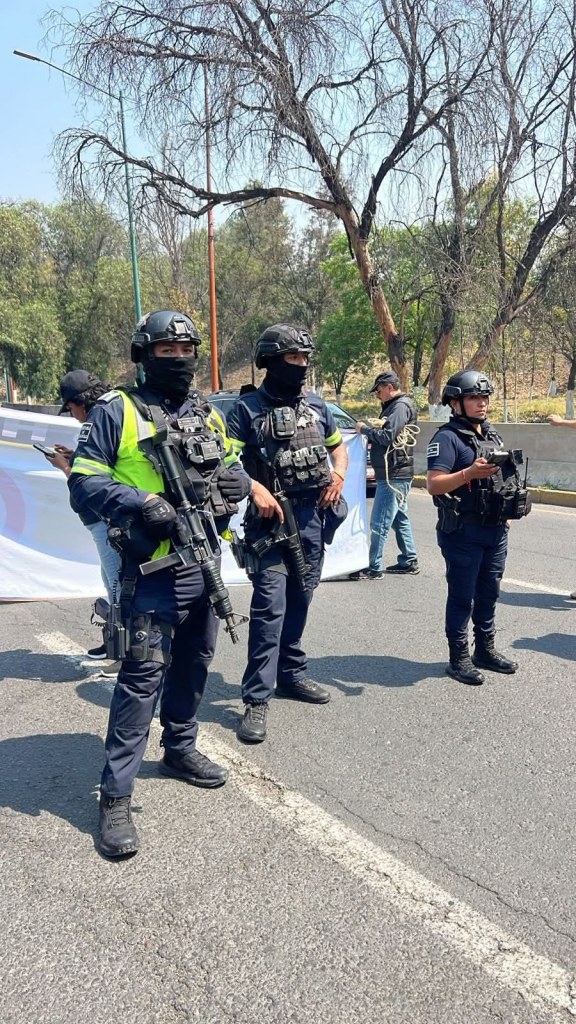 Agentes de policía armados, vestidos con uniforme táctico y máscaras, se encuentran en una carretera durante una protesta, con un fondo de árboles secos.