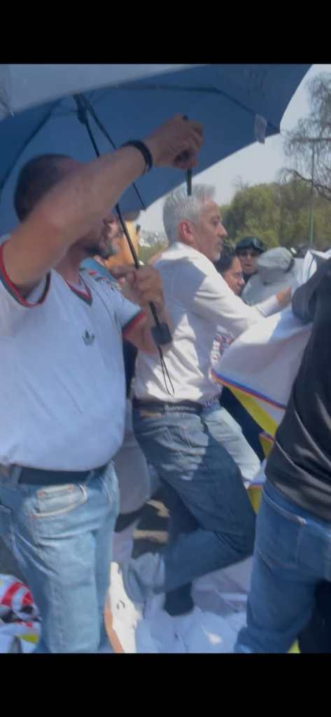 Manifestación con personas sosteniendo un paraguas y pancartas en un ambiente al aire libre.