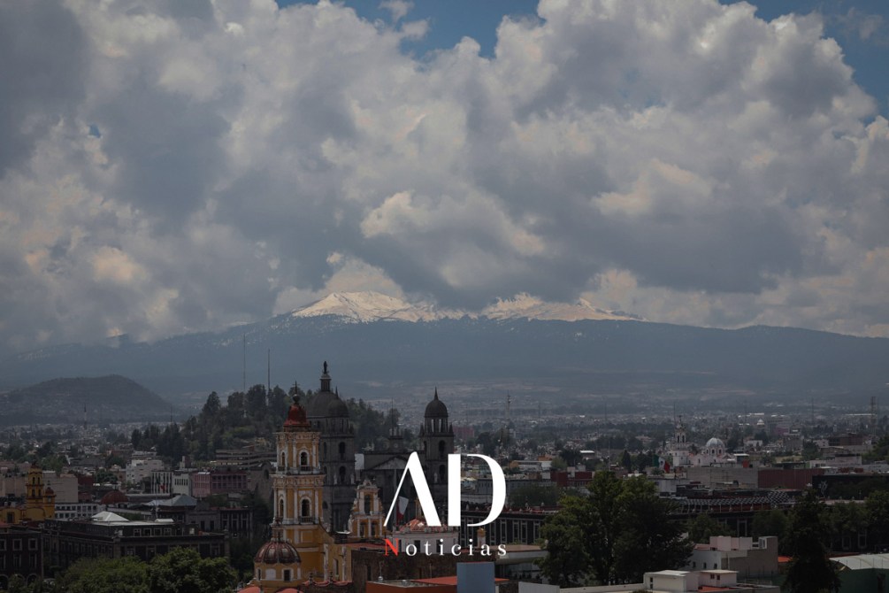 Vista panorámica de una ciudad con edificios arquitectónicos y montañas nevadas de fondo, bajo un cielo nublado.