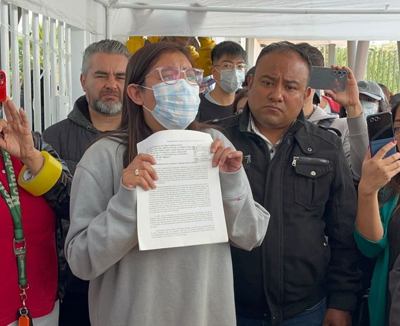 A group of people gathered under a tent, with one young woman in the foreground holding a document, speaking passionately. The crowd includes individuals wearing masks and holding smartphones.