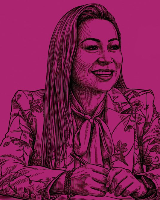 Retrato de una mujer sonriente con un blazer floral y una blusa con lazo, sentada con las manos juntas sobre la mesa, sobre un fondo color púrpura.