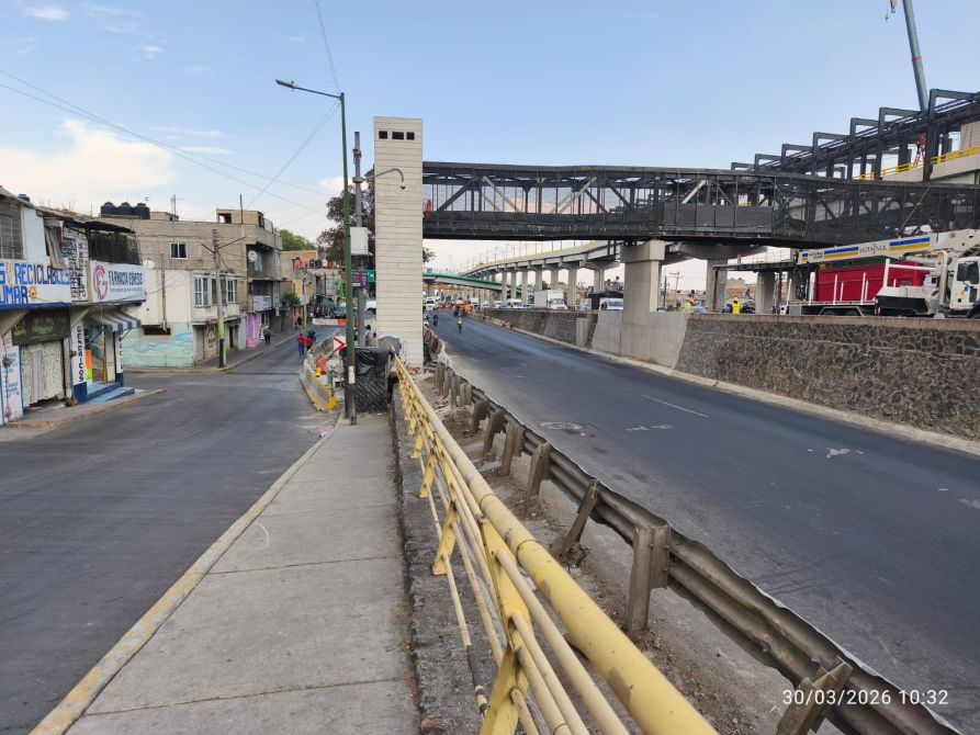 Vista de una calle con una banqueta y una barrera amarilla en el primer plano, edificios a la izquierda y una estructura elevada en el fondo, con un camino pavimentado y vehículos en movimiento.