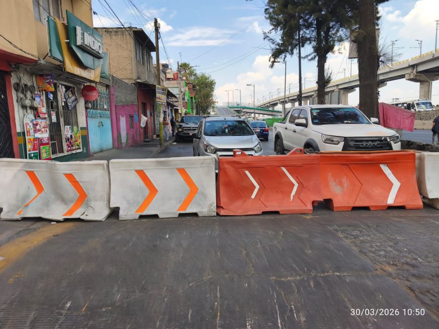 Barricadas de concreto con flechas naranja bloqueando una calle, rodeadas de coches y edificios comerciales.