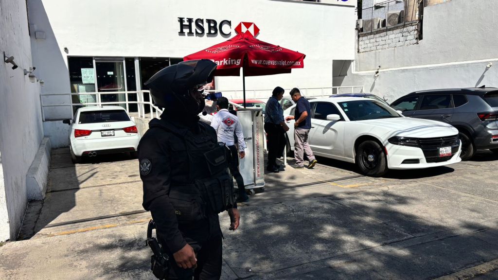 Un oficial de seguridad con uniforme negro se encuentra de pie frente a una sucursal de HSBC, mientras dos hombres conversan al lado de un vehículo blanco y un toldo de valet parking.
