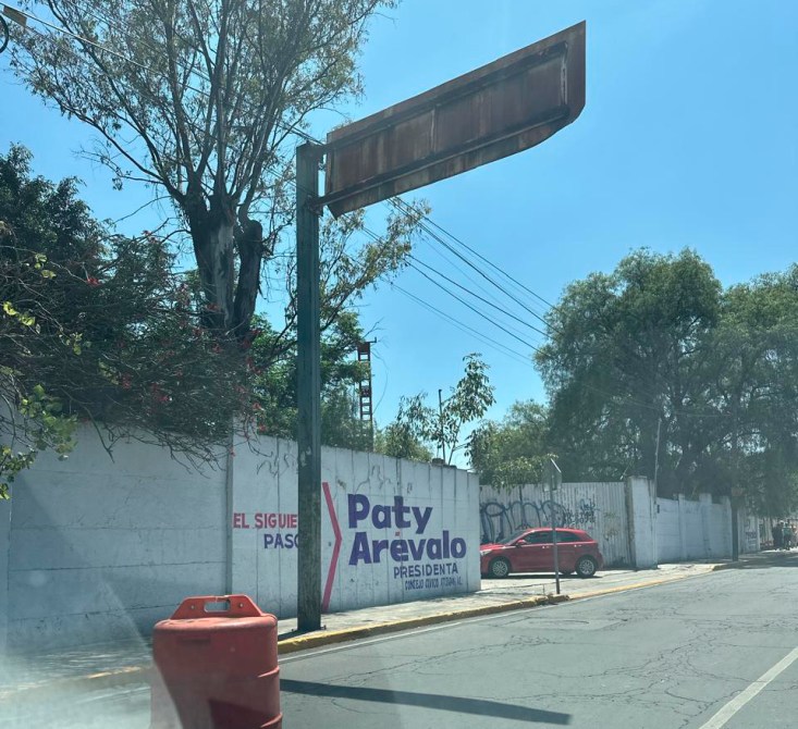 Una señal de tráfico oxidada con un fondo azul claro y un anuncio de campaña electoral de Paty Arevalo para presidenta en una pared blanca. Hay un automóvil rojo estacionado y un tambor de construcción en el primer plano.