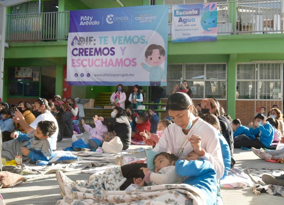 Una madre sostiene a sus hijos pequeños mientras están sentados en el suelo, rodeados de otros niños y adultos en una actividad comunitaria, con un gran cartel que dice 'ADIF: TE VEMOS, CREEMOS Y ESCUCHAMOS'.