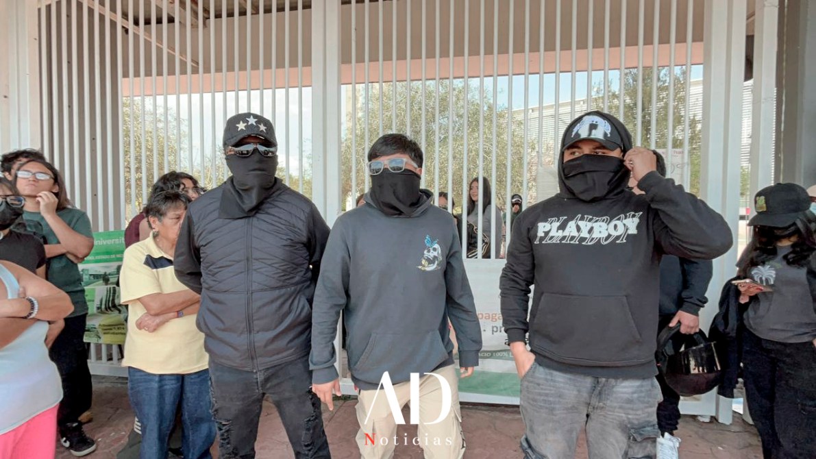 A group of four individuals standing together, wearing black masks and hoodies, with two wearing sunglasses. They are positioned in front of a gated entrance, and several other people are visible in the background.