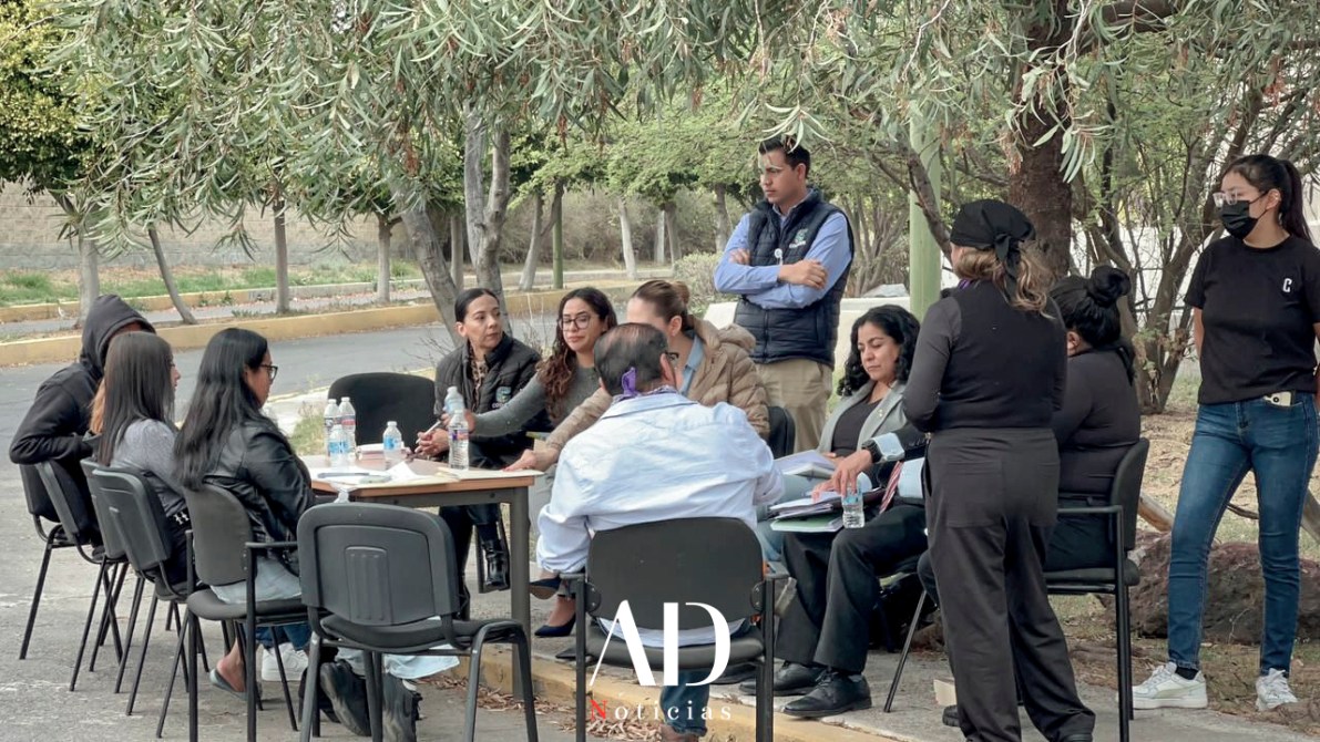 A group of people sitting around a table outdoors, engaged in discussion. Some individuals are taking notes while others are listening. Trees provide shade in the background.