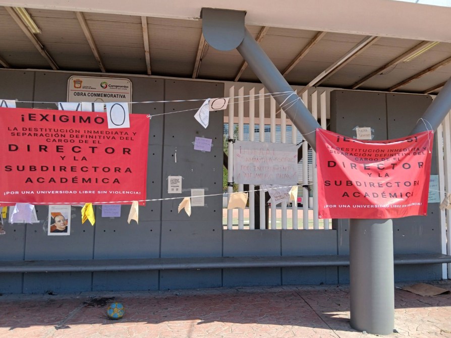 Carteles en una pared exigiendo la destitución del director y subdirectora académica, con letreros en rojo y varios mensajes colgados.