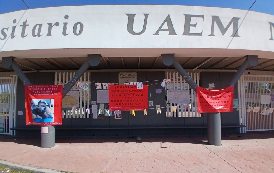 Fachada de la universidad UAEM con pancartas rojas que exigen acción del director y subdirector académico, acompañadas de varios documentos y carteles en las paredes.