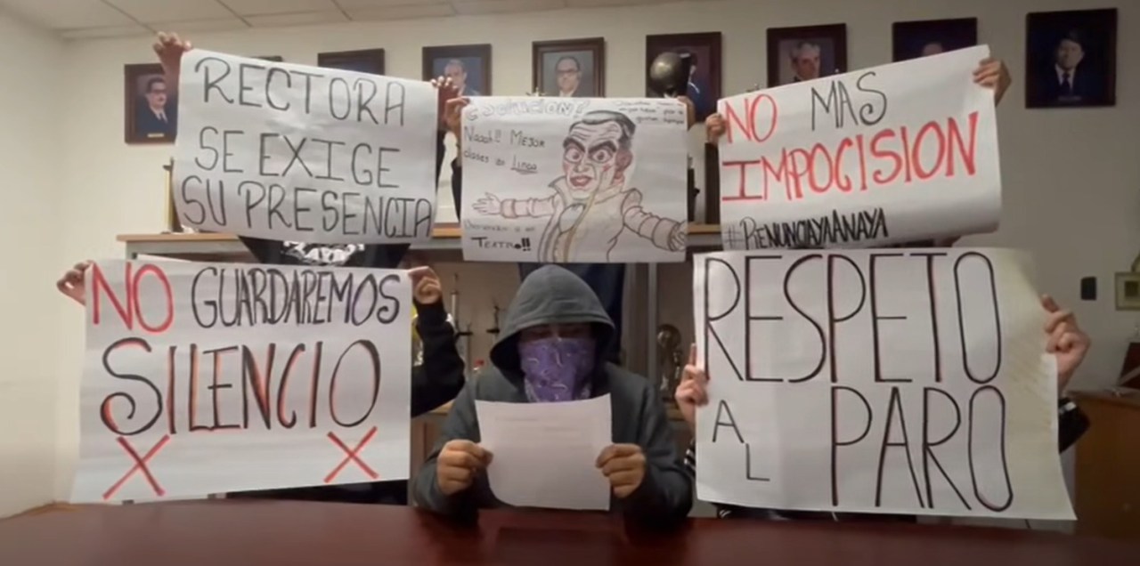 Grupo de personas protestando con pancartas en un ambiente interior, exigiendo la presencia de la rectoría y respeto al paro. Algunos letreros incluyen mensajes como 'No guardaremos silencio' y 'No más imposición'.