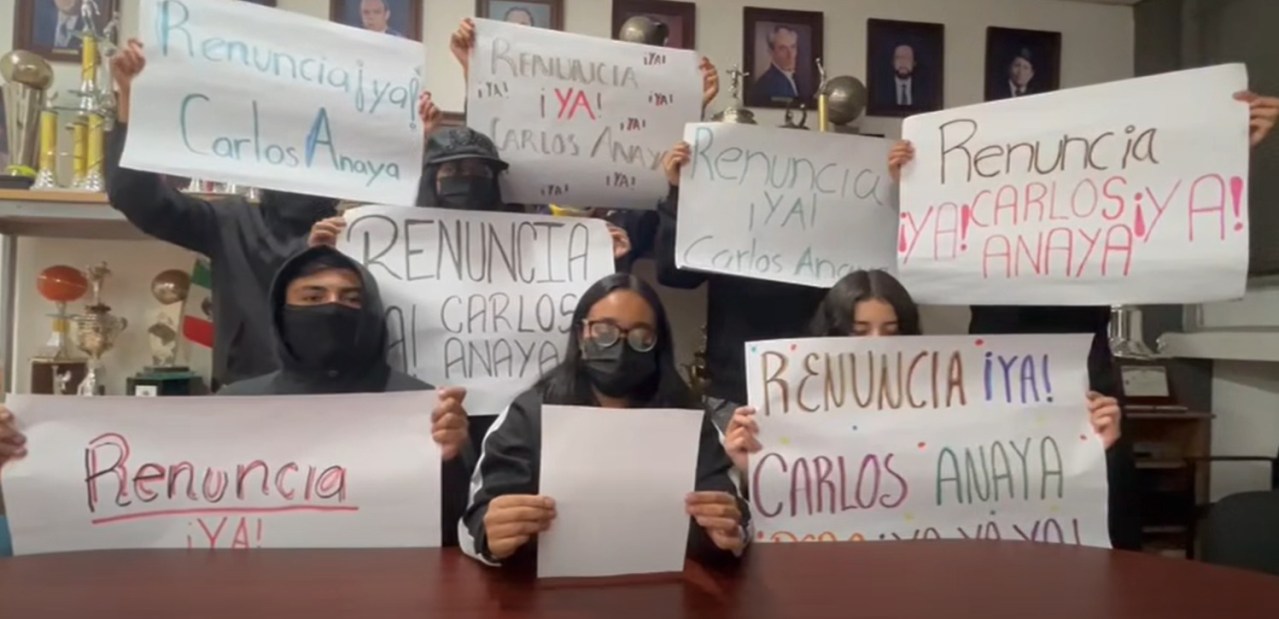 Grupo de personas sosteniendo pancartas que dicen 'Renuncia ya! Carlos Anaya' en un ambiente interior, con trofeos y fotos en el fondo.