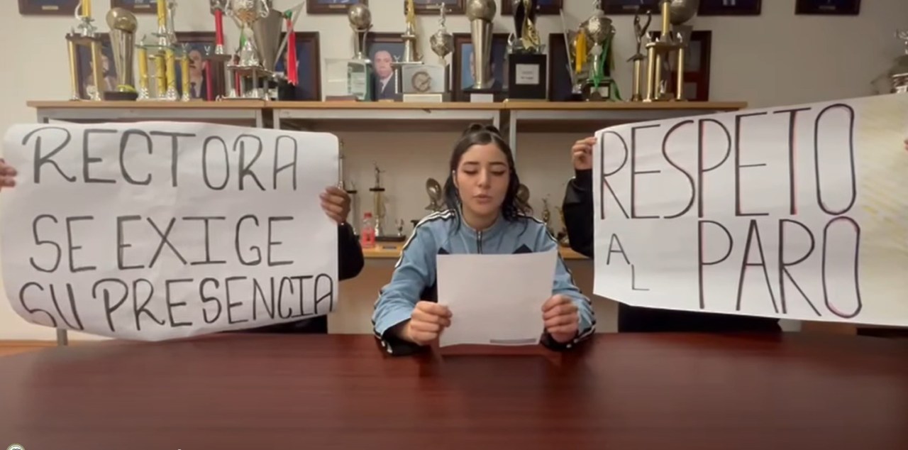 Una joven sentada frente a una mesa lee un documento mientras sostiene una hoja de papel, acompañada de dos personas con pancartas que exigen la presencia de la rectora y respeto al paro.