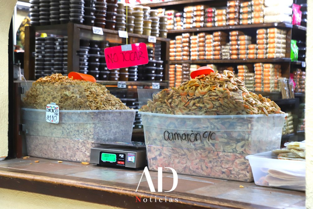 Contenedor transparente con camarones secos a la venta y letrero que dice 'no tocar'. Al fondo, estantes con frascos de productos variados.