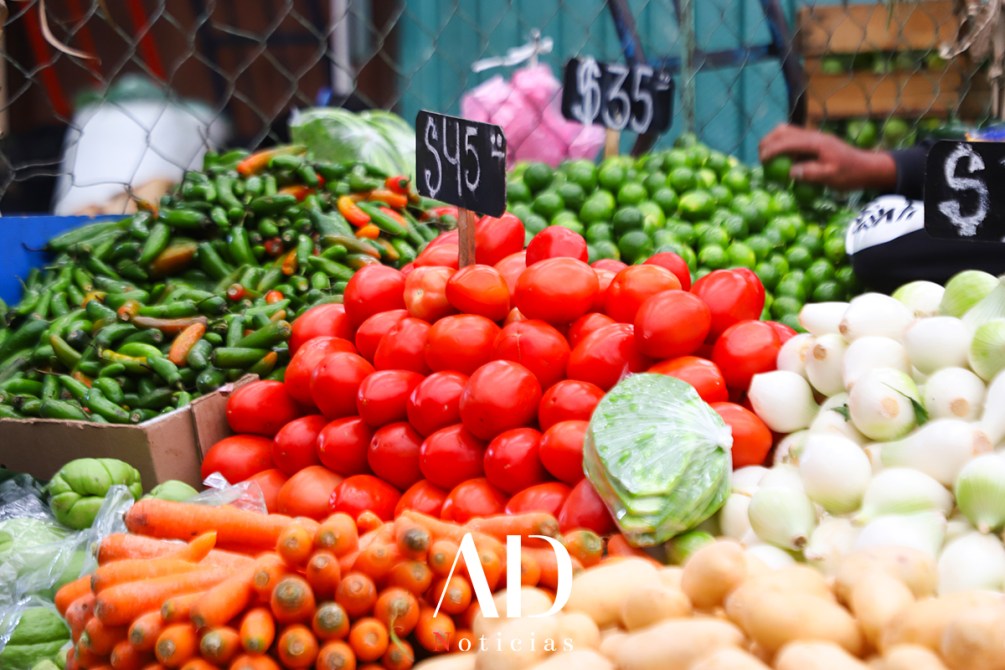 Variedad de verduras y hortalizas en un mercado, incluyendo tomates, chiles, limones y zanahorias, con precios visibles en carteles.