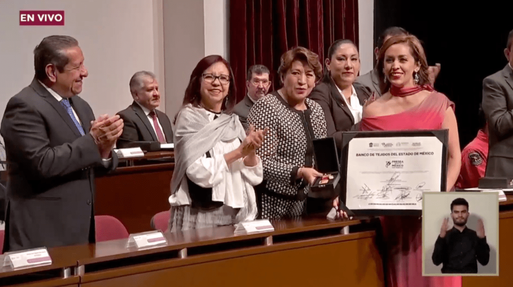 Evento de premiación donde varias personas están aplaudiendo. Una mujer de vestido rojo sostiene un certificado que dice 'Banco de Tejidos del Estado de México'.