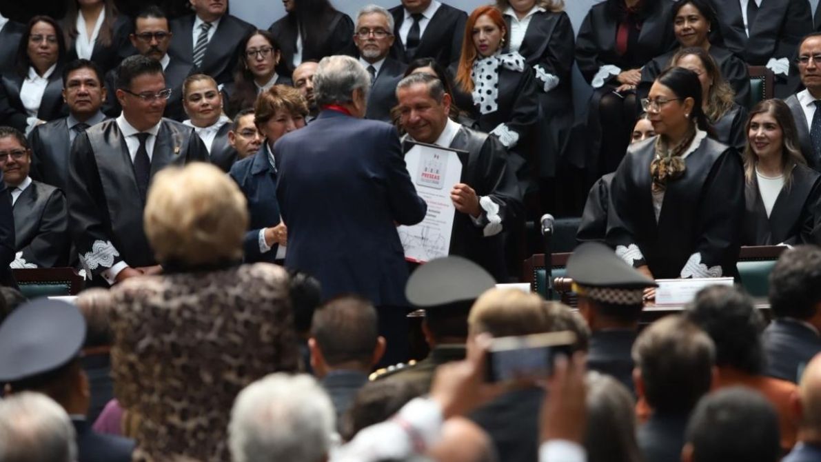 Ceremonia de entrega de un reconocimiento en un congreso, con varias personas en toga observando. Un hombre recibe un documento de manos de otro hombre.