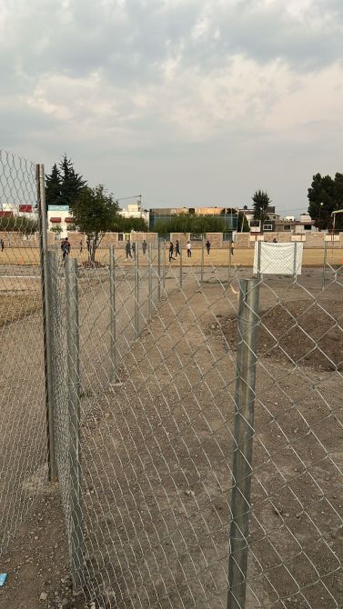A fenced area with a gravel path and a sports goal in the background, where several people are walking on a dirt field.
