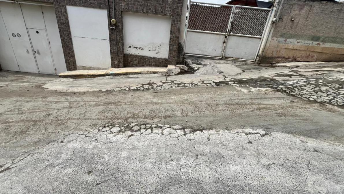 A street view featuring cracked asphalt, a textured wall with garage doors, and a metal gate in the background.