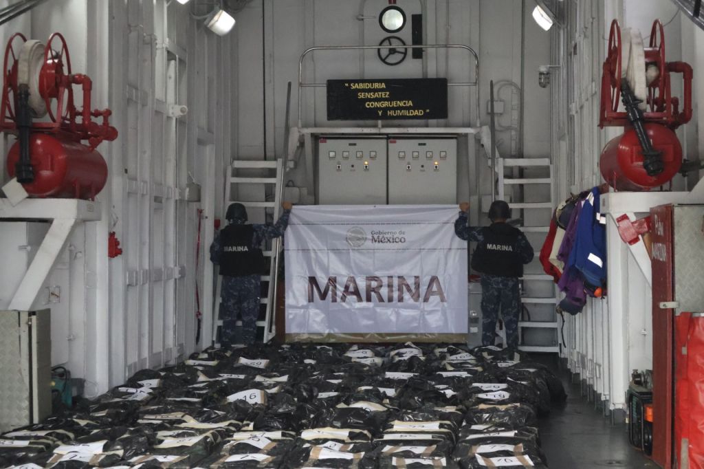 Imagen que muestra a dos miembros de la Marina mexicana sosteniendo una bandera en un espacio con suministros distribuidos por el suelo.