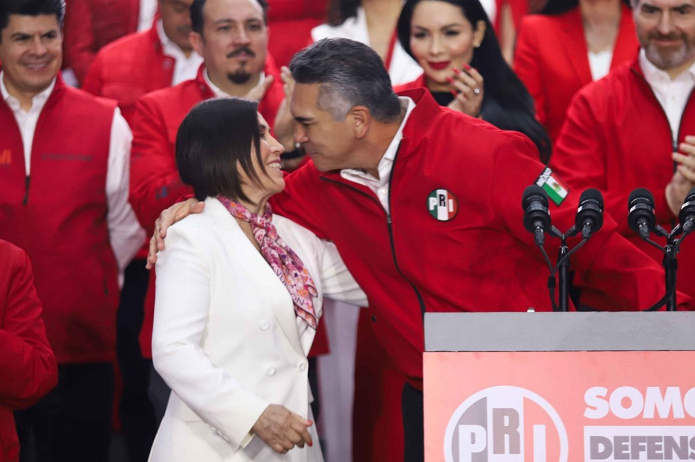Dos personas se abrazan y sonríen en un escenario, rodeadas de otros miembros del partido, todos vistiendo ropa roja. El fondo presenta un cartel del PRI.