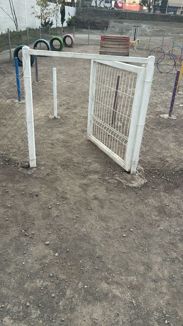 A white gate opening into a fenced playground area with dirt ground and colorful play equipment in the background.