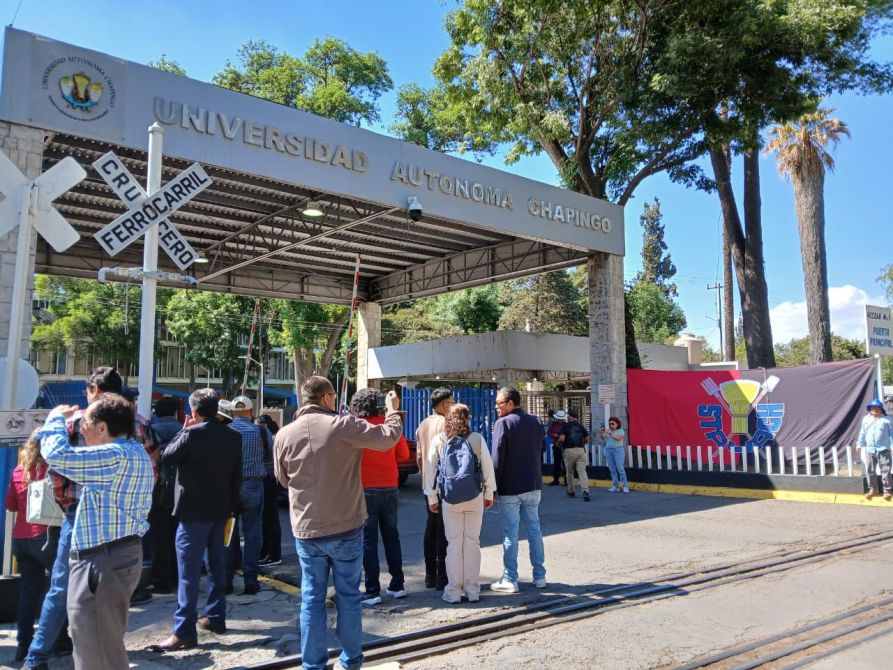 Grupo de personas reunidas frente a la entrada de la Universidad Autónoma Chapingo, con un gran banderín visible y señales ferroviarias en el área.