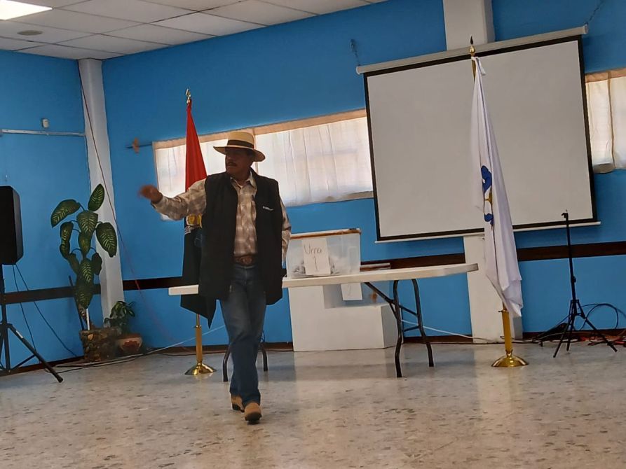 Hombre de pie en un salón con paredes azules, vistiendo gorra y chaleco, presentando frente a una pantalla y una mesa con documentos.