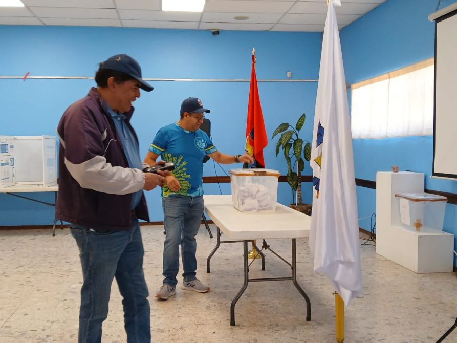 Dos hombres en un lugar de votación, uno sostiene un dispositivo mientras se acerca a una urna con boletas. Hay banderas y una planta decorativa en el entorno.