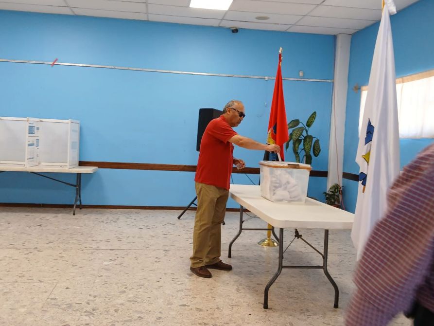 Hombre depositando su voto en una urna en un salón de votación con bandera roja y blanca.