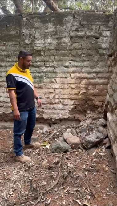 A man standing in a sparse area with a cracked brick wall, observing the ground covered with stones and dry leaves.