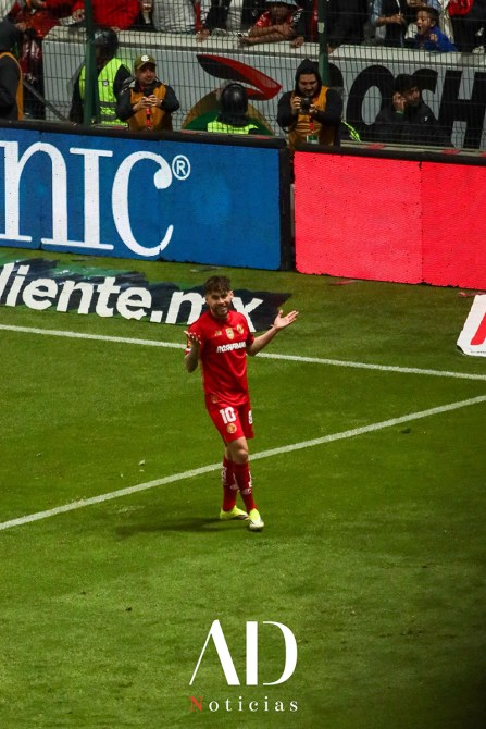 Jugador de fútbol celebrando en la cancha, vestido con camiseta roja con el número 10, rodeado de aficionados en un estadio.