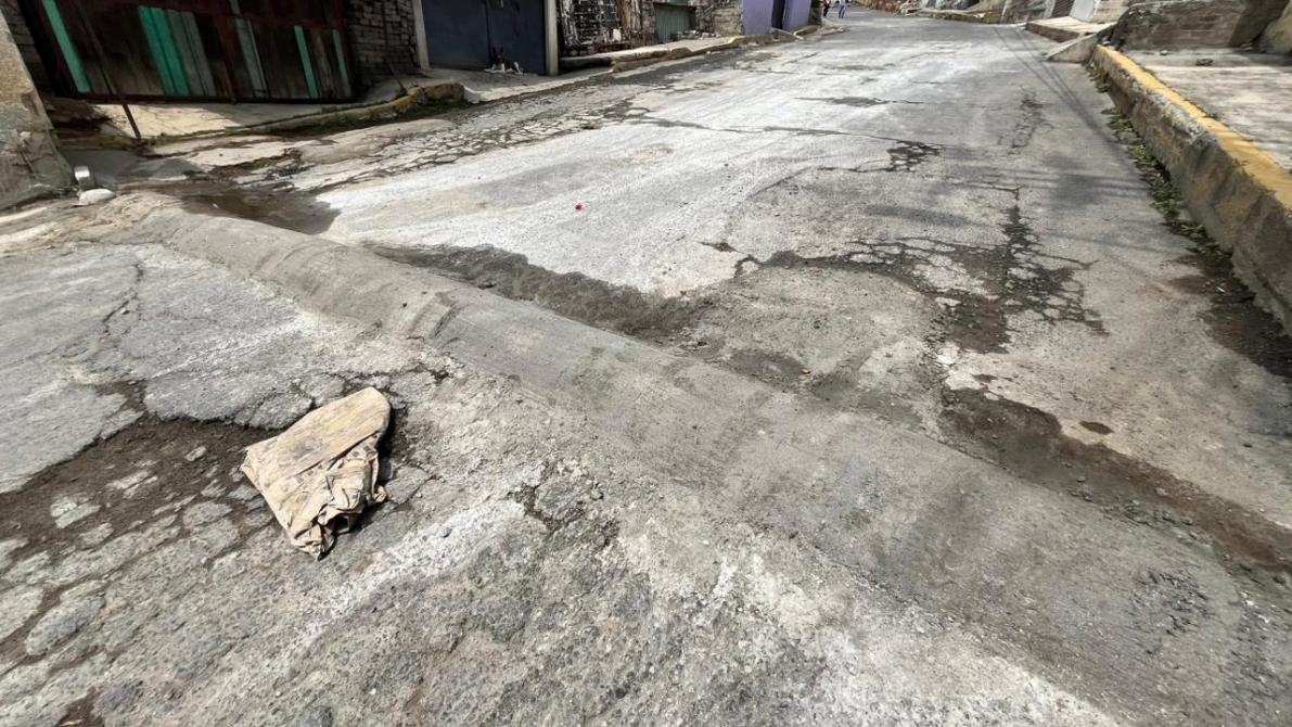 A cracked and uneven street showing patches of asphalt and debris, including a piece of fabric, with buildings in the background.
