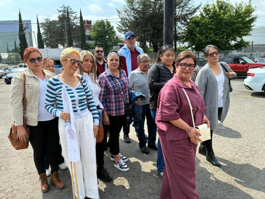 Grupo de personas alineadas en un lugar al aire libre, sonriendo y posando para la cámara. Algunos llevan gafas de sol y vestimenta casual. Se observan árboles y edificios al fondo.