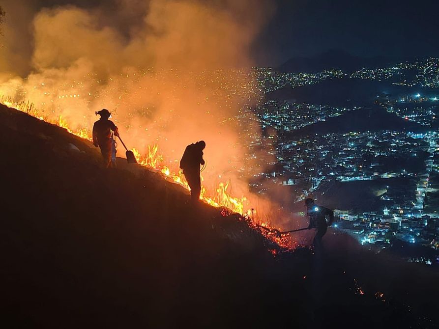 Bomberos combatiendo un incendio forestal en la ladera de una montaña durante la noche, con luces de la ciudad al fondo.