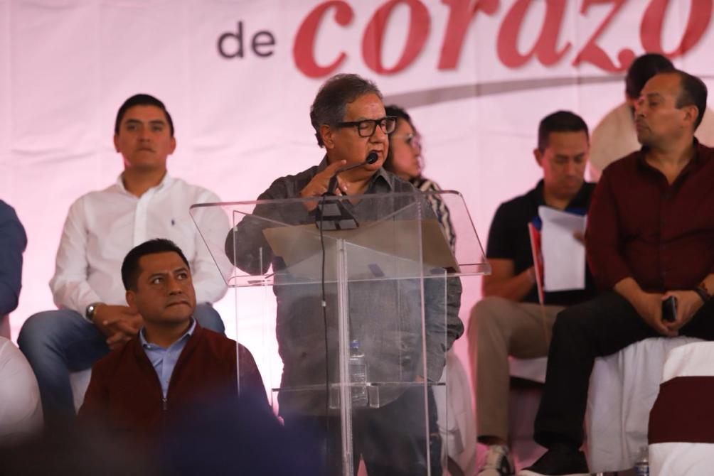 Un orador en un podio durante un evento, con un mensaje de fondo que dice 'de corazón'. Varios asistentes observan atentamente.