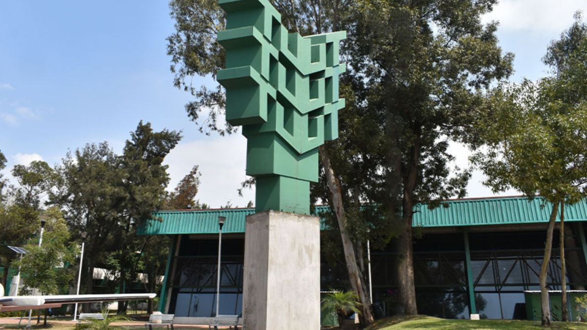 Escultura abstracta de color verde ubicada en un parque, con un fondo de edificios y árboles.