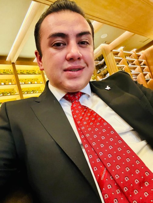 Hombre joven con traje negro y corbata roja con patrones, sonriendo en un entorno elegante.