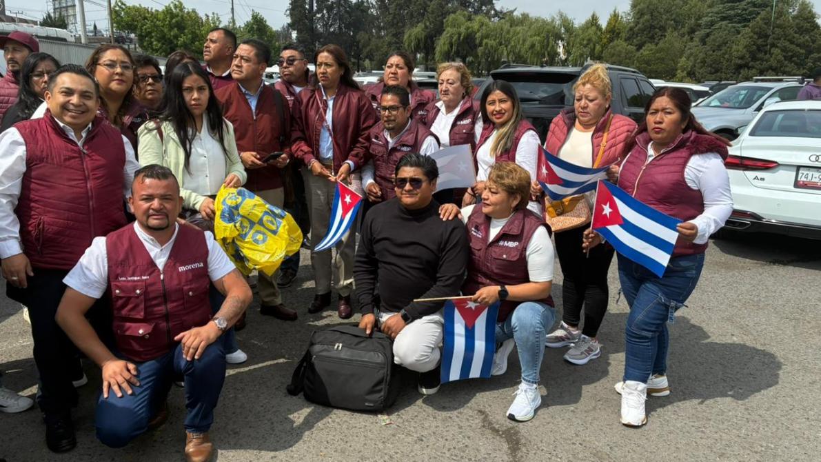 Grupo de personas reunidas, algunas sosteniendo banderas cubanas, con chaquetas de color burdeos y sonrisas, en un ambiente al aire libre.