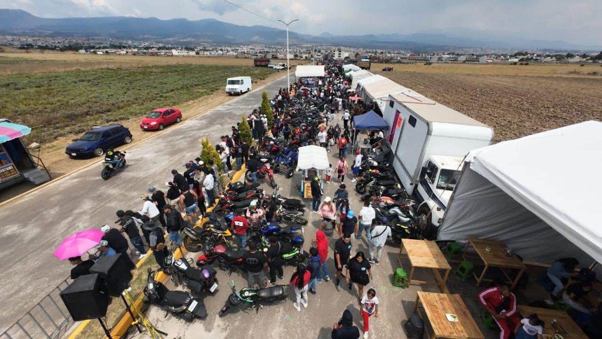 Vista aérea de un evento con numerosas motocicletas y personas en un área rural, con camiones y carpas en los lados.