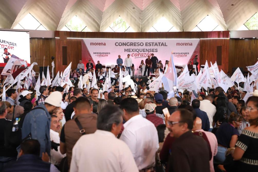 Evento masivo del partido Morena en un congreso estatal, con una multitud de personas sosteniendo banderas y pancartas dentro de un recinto iluminado.