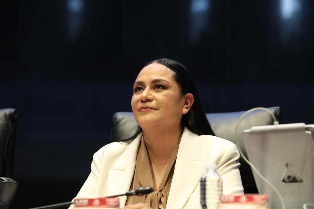 Mujer con cabello largo y oscuro, vestida con un blazer blanco, sentada en un entorno de reunión, mirando hacia arriba con una expresión contemplativa.